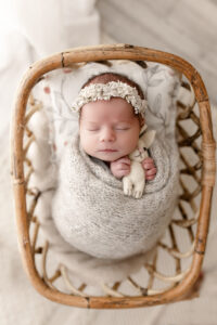 Newborn posed in tiny wicker bed, wrapped in soft knit wrap holding a felt bunny