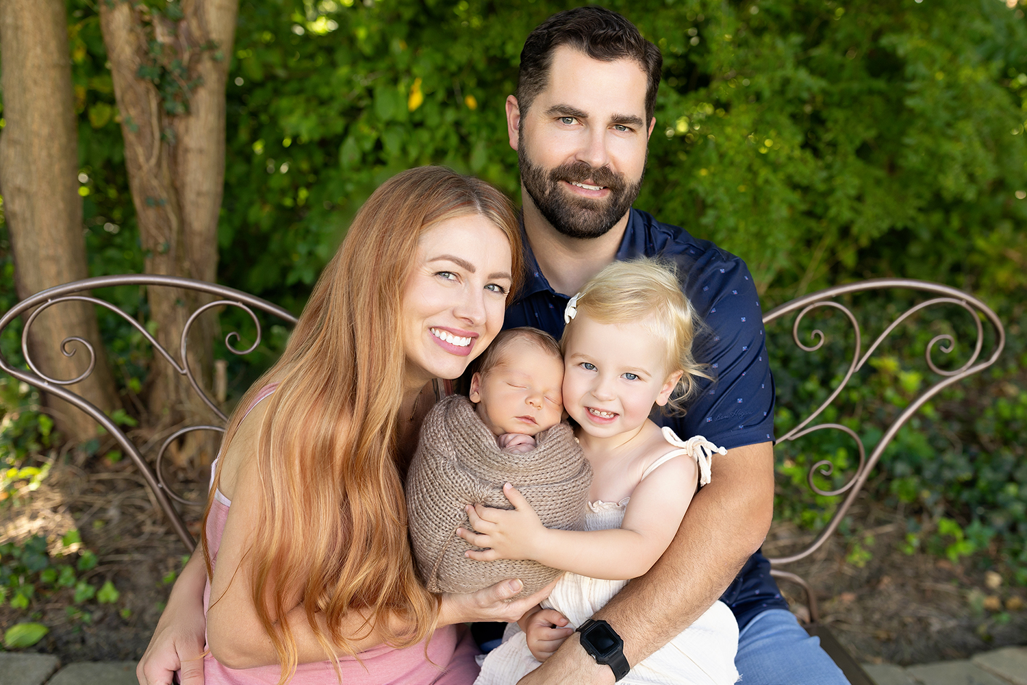 A family of four cuddling closely outdoors during a newborn session, with the parents lovingly holding their swaddled newborn while their older child snuggles beside them, all surrounded by soft natural light and a peaceful outdoor setting.