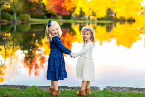 Two sisters hold hands in front of a pond with fall colors reflecting in the water. 