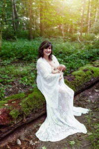 Mom smiles at the camera as she nurses her newborn baby sitting on a tree with beautiful moss growing all over it. 