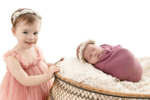 Big sister barefoot and wearing soft muted tones posing with baby sister during family newborn session